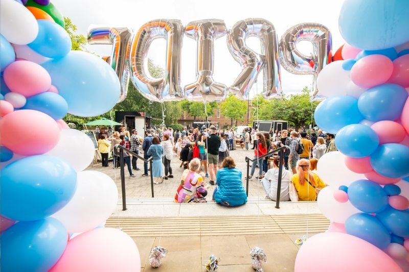 Image of lgbt and lgbt ally community attending the Pride Picnic event outside the Elmwood hall on a cloudy day with a large set of silver letter balloons spelling 'Pride'